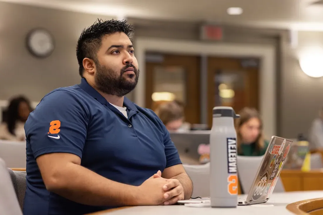 Man sitting at desk in classroom with laptop and water bottle.